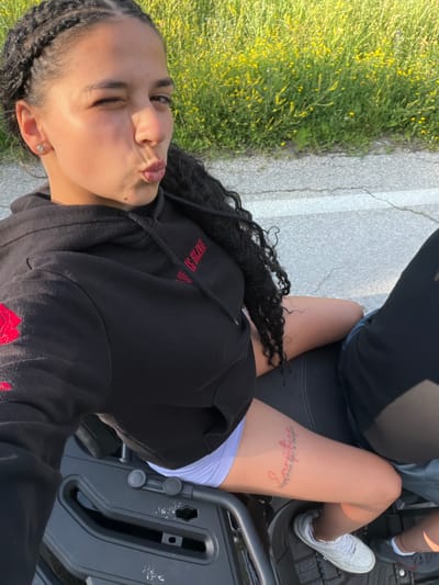 In a vertical, eye-level shot taken outdoors in Boltata, Bulgaria, a young woman is captured mid-pout, her eyes squeezed shut. She's wearing a black hooded sweatshirt with red embroidery and white shorts. A red tattoo on her thigh reads "Everything happens for a reason."

The setting appears to be a roadside, with a cracked asphalt surface and a grassy verge dotted with yellow wildflowers. The sun is high, suggesting it's daytime, and the weather is clear and bright. A dark grey, possibly plastic, seat is visible, hinting that the woman might be on a vehicle like an ATV or motorcycle. The scene conveys a sense of youthful exuberance and a relaxed outdoor moment.