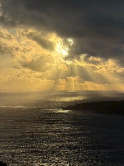 The image displays a panoramic coastal landscape, identified as being in Sakti, Indonesia. The upper half of the frame is occupied by a dramatic sky featuring heavy, dark grey clouds. A bright, obscured sun emits numerous distinct golden crepuscular rays that penetrate the clouds and extend downwards, illuminating the upper-central and upper-left portions of the sky with intense golden light. Below the horizon, a vast expanse of dark, choppy ocean water fills the mid-ground and foreground, reflecting pockets of the golden sunlight on its surface. To the lower-right, a dark, silhouetted landmass, appearing as a vegetated coastline or island, extends into the water, forming a natural curve or cove. A faint, distant landmass or atmospheric haze is visible along the far horizon line. No people, structures, or explicit actions are discernible.