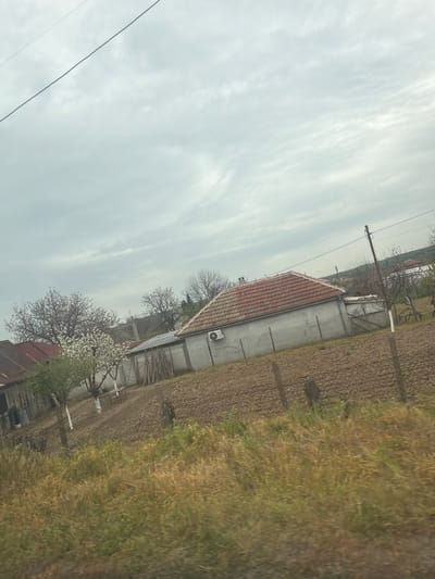 The image depicts a rural scene in Seltsi, Bulgaria, featuring a one-story building with a red-tiled roof and light-colored walls. An air conditioning unit is attached to the exterior wall. Adjacent to the building is a field with dry, brown vegetation. Trees, some blossoming, are interspersed near the building and in the distance. Above the scene is an overcast sky. Power lines are visible in the upper left corner.