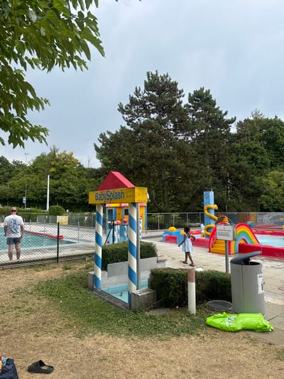 In Renens, Switzerland, at a public swimming pool facility, an overcast day is depicted. In the foreground, dry, patchy grass is visible, with a black sandal and a green inflatable object lying on the ground. A large grey trash bin is situated to the right. Behind this, a child in a light blue dress is walking in a shallow play pool area featuring a rainbow slide and water features. Further to the left, a man wearing a patterned white t-shirt and grey shorts stands facing the play area. He appears to be on the edge of a larger swimming pool, separated by a fence. The background is filled with lush green trees and a cloudy sky. A sign on the play area reads "BabySplash MAX 5 ANS".