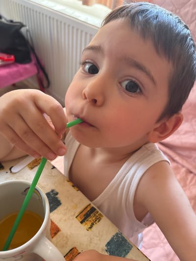 An indoor domestic setting in Barakovo, Bulgaria, features a young child, approximately 2-4 years old, with fair skin, dark brown hair cut in a short style with a fringe, and large dark eyes. The child is wearing a white ribbed sleeveless undershirt and is positioned at a table, looking directly at the camera with a wide-eyed expression. The child is actively drinking from a bright green plastic straw, which is inserted into a white ceramic mug containing an orange liquid, likely juice. The mug features a small, faded rainbow decal. A light-colored patterned tablecloth covers the table surface, displaying abstract rectangular and square designs in hues of orange, blue, and black. A portion of a white spoon handle is visible on the tablecloth. In the background to the left, a white horizontal-finned radiator is prominent. Below the radiator, a dark-colored bag rests on a pink fabric item. The immediate background to the child's right and behind the child is a soft pink textile surface, resembling a blanket or bed sheet. The scene is brightly illuminated, suggesting natural light from an unseen window.