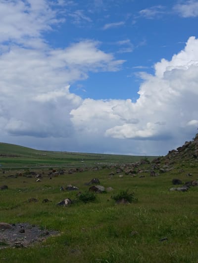 The image shows a landscape near Tlik, Armenia. The left side features a blue sky with white cumulus clouds. The right side shows a green hillside with scattered rocks. In the middle ground, there is a flat expanse of green land. The image appears to have been taken from a high vantage point, possibly overlooking the terrain. There are no people visible in the frame. The main subjects are the sky, clouds, hillside, and grassy plain. The environment is rural and mountainous. The action is stillness, a static scene.