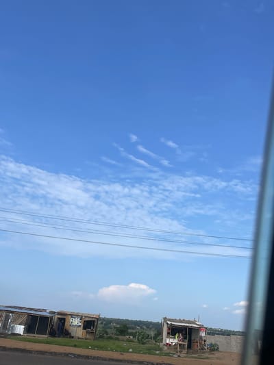 This image, captured in Uke, Nigeria, depicts a daytime roadside scene under a clear blue sky with scattered cumulus clouds. Multiple dark power lines stretch horizontally across the middle ground.
On the left, two adjacent, open-fronted, single-story structures with corrugated metal roofs and wooden frames are visible. The structure on the far left appears unfinished or in disrepair, with visible wooden scaffolding and a partial corrugated metal roof. The structure immediately to its right is more complete, featuring walls that appear to be a mix of wood planks and possibly sheet metal. Several individuals are present in and around these structures; at least two people are seated outside the second structure, and others are partially obscured inside. Signs with text and images are affixed to the front of this structure.
Further to the right, a third, smaller, single-story roadside stall is visible. This stall also has a corrugated metal roof and appears to be constructed from wooden planks. It features an open counter area where goods are displayed, and at least one person is visible behind the counter. A red sign with white text, partially reading "POS," is attached to the front right side of this stall.
The foreground consists of a dirt and gravel road shoulder with sparse green vegetation and scattered debris. Beyond the structures, the landscape recedes into rolling hills covered with greenery under the distant sky. A concrete or stone wall is partially visible behind the rightmost stall. A dark vertical element on the far right edge of the frame suggests the image was taken from inside a vehicle, possibly through a window frame.