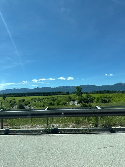 The image depicts a scenic landscape under a clear blue sky with scattered white clouds. In the foreground, a highway guardrail runs horizontally across the frame, separating the road from a grassy field. The field is lush green, dotted with various shrubs and trees, and in the distance, a range of dark, wooded mountains stretches across the horizon. The overall impression is one of a vast, open natural environment. The image does not provide any specific details about the city of Barlete, Croatia.