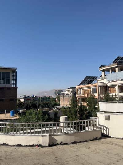 This is an eye-level outdoor photograph taken on a clear day in Kabul, Afghanistan. The foreground shows a concrete rooftop with a metal railing. Beyond the rooftop, a dense cluster of trees forms a green barrier, partially obscuring a collection of buildings. The buildings vary in architectural style and color, with some featuring balconies, large windows, and solar panels installed on their roofs. In the distance, a mountain range is visible under a hazy sky. One building on the left has a brick base and a modern glass structure on top, and a blue water tank is visible near it. Several vehicles and what appear to be smaller residential structures are also interspersed within the urban landscape. The overall impression is of a modernizing city with a mix of traditional and contemporary elements, set against a backdrop of natural landscape.