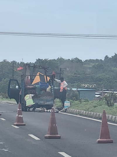 In Ghodasgaon, India, on what appears to be a highway, a blue truck is parked with its rear open, loaded with various items including sand, a blue container, a generator, and some orange construction cones. Two individuals are present. One person, wearing a high-visibility yellow vest and dark pants, is positioned in the back of the truck, seemingly attending to the cargo. Another person, wearing an orange vest over a white shirt and dark pants, stands next to the truck, facing away from the camera. The road is marked with white lines and several orange traffic cones are placed along the side, indicating a work zone. In the background, there are trees and a small building structure, under a cloudy sky.