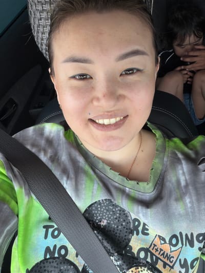 The image shows a woman in the driver's seat of a car, with a child in the back seat. The woman is wearing a tie-dye t-shirt and a grey headband. The child is wearing a black shirt. The car appears to be moving, as indicated by the motion blur in the background. The scene is consistent with a person driving in the city of Dams, Kazakhstan.