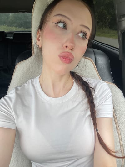 A young woman with light skin, dark brown hair braided to her right, and green eyes is sitting in the driver's seat of a car. She is wearing a white t-shirt. She is looking to her right and her lips are slightly pursed. The interior of the car is visible, including the headrest and a fluffy white seat cover. The background outside the car windows shows trees and foliage, suggesting a rural or suburban setting. There are no discernible landmarks to indicate the specific location within Gavrilovka, Russia.