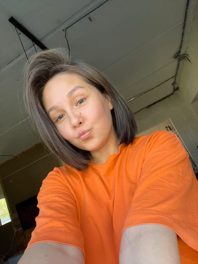 A selfie of a woman with short grey/brown hair wearing an orange t-shirt. The background suggests an interior with bare walls and exposed electrical wiring. The location is claimed to be Verkh-Katunskoye, Russia, but there are no elements within the photograph that can confirm this.