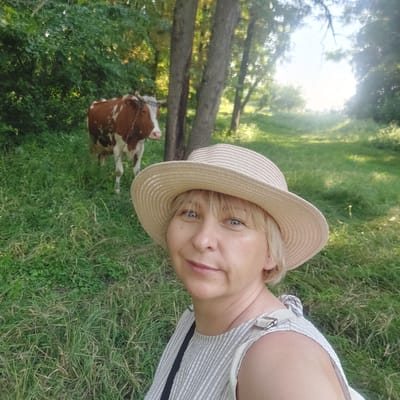 The image is a selfie taken outdoors in a grassy, wooded area, possibly in Voronkiv, Ukraine. The primary subject is a woman wearing a straw hat and a striped dress. She is smiling at the camera, with her face partially obscured by the brim of her hat. Her blue eyes are visible, and she has blonde hair.

In the background, a cow with brown and white markings is grazing or standing in the grass. Several trees are also visible, with sunlight filtering through the leaves, suggesting it is daytime. The overall scene appears to be a peaceful, rural setting. The woman's attire and the presence of the cow indicate a casual, perhaps rural or agricultural, environment. There is no visible text in the image.