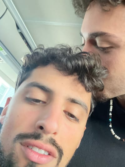 The image depicts two males inside a vehicle, likely in transit. The male in the foreground, with dark curly hair, a mustache, and goatee, looks toward the viewer. The second male, visible behind and to the right, has his eyes closed and is pressing his lips to the forehead/hair of the foreground male. He wears a necklace composed of white, black, and turquoise beads. The upper left of the frame shows the vehicle's interior paneling and a blurry external view of a light-colored building with siding. The specific location within New Brunswick, United States, is not identifiable from the visual information provided.