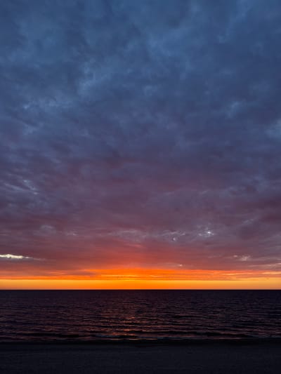 The image depicts a sunset over the Baltic Sea, near Garupe, Latvia. The sky exhibits a gradation from dark blue with textured clouds on the left, transitioning to orange and yellow hues towards the horizon. Below the horizon, the ocean surface reflects the sunset colors. A narrow strip of dark sand is visible at the bottom right of the frame. No people or identifiable objects are present.