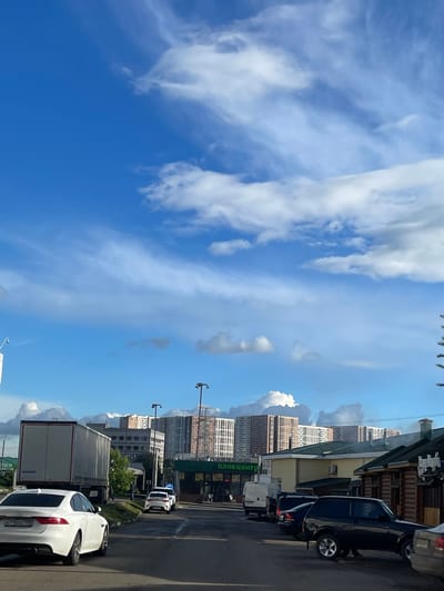 The image captures an urban scene in Bittsa, Russia. A clear blue sky with wispy clouds dominates the left side of the frame. On the right, buildings and various vehicles line a street. These vehicles include cars and a truck. The buildings appear to be a mix of residential and commercial structures. Street lights are also visible along the road.