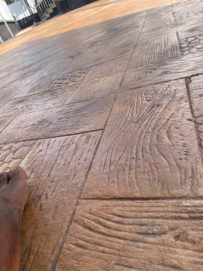 A low-angle, close-up view depicts a dark-skinned bare human foot, with five toes visible, positioned at the bottom-left of the frame. The foot rests on an outdoor paved surface composed of reddish-brown and dark brown stamped concrete or tiles. The paving features a repeating pattern primarily mimicking wood grain in rectangular sections, interspersed with square sections exhibiting a raised cobblestone or pebble-like texture. The surface appears dry with minimal debris. In the blurred background, partial structures are visible, including white and dark building elements and a metallic railing, under daylight illumination. The scene is located in Ogbunu, Nigeria.