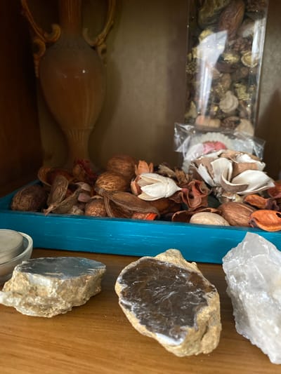 The image displays a close-up, eye-level view of a shelf in Osek, Czechia, showcasing a collection of decorative items. In the background, a light brown, ornate vase with gold accents stands to the left, partially obscured by shadow. To its right, a clear plastic container filled with potpourri, featuring various dried botanicals and seed pods, is positioned behind a small, clear plastic bag containing a white, ruffled object with pink accents.

The foreground is dominated by a shallow, turquoise-colored rectangular tray filled with more potpourri, consisting of a variety of dried leaves, seed pods, and floral elements in earthy tones of brown, red, and beige. In front of the tray, resting on a wooden surface, are three distinct geological specimens. The leftmost is a flat, grayish-blue stone with visible crystalline structures and a rough, light brown edge. To its right is a darker, brownish-black stone, also flat and featuring white, vein-like inclusions. To the right of this, a larger, translucent white crystalline rock stands upright. The lighting is soft, with shadows suggesting an indoor environment. No individuals are present in the scene.
