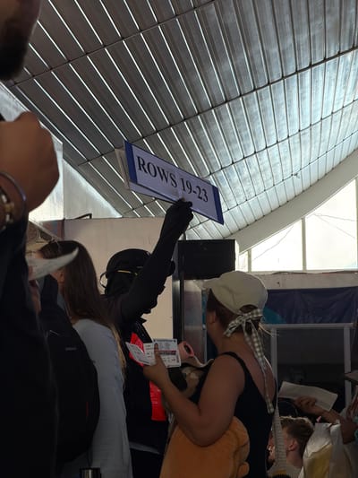 The image shows an indoor scene, likely a seating area or waiting room, potentially in Del Carmen, Philippines. People are present, with a woman wearing a cap and black tank top. Another individual is partially visible, wearing black clothing and potentially a backpack. Someone is holding a sign displaying "ROWS 19-23". The setting features what appears to be a slatted wall or ceiling structure. The overall scene suggests a process of checking or directing people to specific rows, possibly at an event or transportation hub.