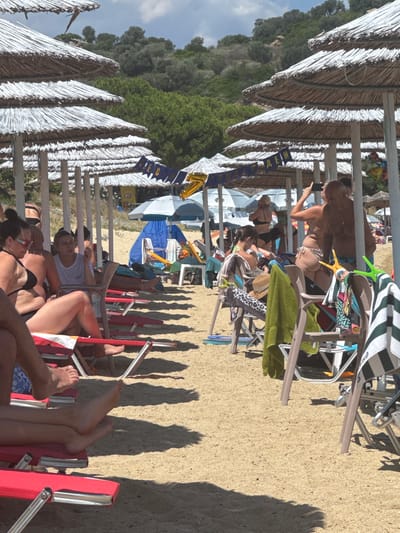 The image depicts a crowded beach in Ammouliani, Greece, with numerous thatched umbrellas providing shade over rows of beach chairs on the sand. Several people are visible, most of whom are seated on the chairs, some wearing swimwear. In the background, a lush green hillside dotted with trees rises behind the beach. A blue tent is set up further back. A banner with yellow lettering and a gold balloon shaped like the number "7" are suspended between some of the umbrella poles, suggesting a celebration or event. The scene is characteristic of a busy summer day at a popular tourist destination.