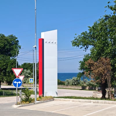 In the city of Pervomaiske settlement, Ukraine, on a clear, sunny day, a modern, tall, rectangular structure with a red vertical stripe on its left side stands prominently. This structure is flanked by a metal pole with a security camera and a street lamp. To the left of the structure, a series of traffic signs are visible: a large, white, inverted red triangle indicating yield, a smaller similar sign below it, and a blue circular sign with two opposing arrows, signifying a choice between turning left or right. A yellow square sign with a pedestrian symbol is also present. In the foreground, a paved parking lot with white lines is delineated by a concrete curb. Beyond the parking lot, a road stretches into the background, separated by a low metal fence and a line of small green and yellow flowering plants. Lush green trees border the scene on the left and right, with a glimpse of the blue sea visible in the distance between the trees and the road. The environment suggests a public space, possibly near a coastal area, with a focus on traffic management and modern urban design.