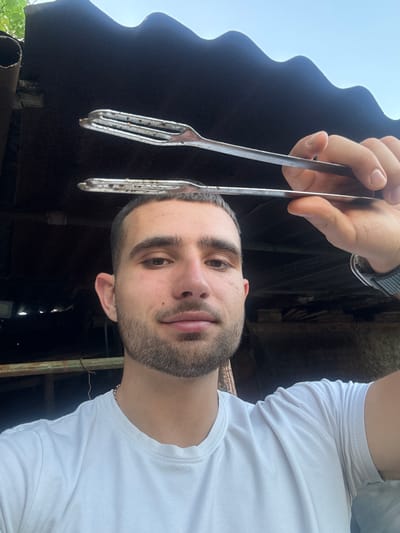A man in a white t-shirt holds a pair of metal barbecue forks. The man has dark hair and a beard. He wears a watch on his left wrist and a small chain around his neck. The background is an outdoor, rural environment, potentially Boltata, Bulgaria, with a dark roof-like structure and hints of greenery.