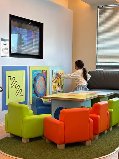 The image displays an indoor children's play or waiting area located in Kiryat Ono, Israel. A young girl, viewed from the side-rear, with dark, wavy hair tied back, is wearing a light-colored long-sleeve top and a grey skirt. She is leaning forward, interacting with the rightmost of three vertical, rectangular, wall-mounted panels designed for play, manipulating a small knob or latch on its light brown, house-themed surface. The adjacent middle panel features a circular, multi-colored spiral, and the leftmost panel shows a black, wavy maze pattern on a light green background. Above these panels, a black-framed flat-screen television displays what appears to be a news program with two male figures and visible Hebrew text. To the left of the television, a sign in Hebrew and English indicates "THIS PLACE IS PROTECTED BY CCTV CAMERAS." The floor is covered by a circular green rug, upon which several child-sized chairs in bright lime green and orange are arranged, alongside partially visible blue chairs. Two low, child-height tables with light blue bases and colorful, decorated tops are positioned between the girl and the foreground chairs. A dark grey upholstered sofa is visible along the right wall. A large window with horizontal venetian blinds is situated on the far right, admitting natural light into the space, which has light-colored walls and ceiling with integrated lighting.