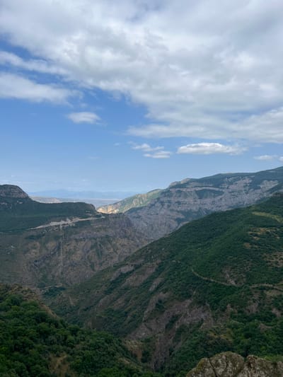 The image displays a panoramic view of the Vorotan River Gorge in Tatev, Syunik Province, Armenia. The scene is dominated by a deep, rugged gorge featuring steep mountain slopes. These slopes are heavily covered in dark green vegetation, interspersed with visible sections of grey and brown rock. A light-colored, winding road or track is discernible traversing a forested slope on the left side of the gorge. In the background, distant mountains appear hazy, with a distinct sunlit area illuminating a segment of the valley floor. The sky above is blue with scattered white cumulus clouds. Dark, angular rock outcrops are present in the immediate foreground on the lower right. No human or animal figures are visible within the frame.