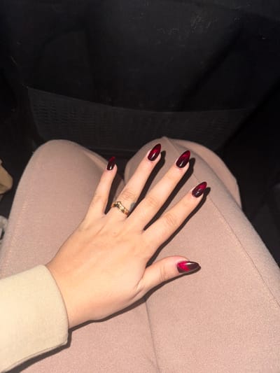 A woman's hand rests on a pair of tan-colored pants, possibly in a car. Her nails are almond-shaped with a dark, red-to-black ombre design, giving a striking appearance. A gold-colored ring adorns her ring finger. The background is partially shadowed, with a hint of the car's interior visible. The sleeve of her beige-toned top is also visible.