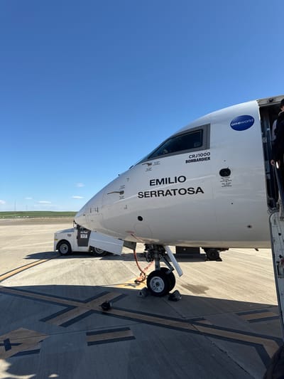 Here's a description of the image:

The image shows a Bombardier CRJ1000 aircraft named "EMILIO SERRATOSA" bearing the "oneworld" logo. The airplane is likely parked on a tarmac, possibly at the airport in Alguaire, Spain, given the reference to its location. A small AirRail 117 baggage tow vehicle is visible near the front landing gear of the plane. The tarmac is marked with striped lines. The sky is clear and blue. There are some personnel in the doorway.