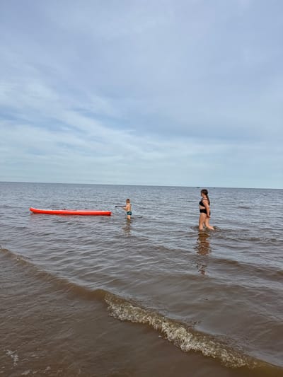 This is a vertical, full-shot photograph taken from a high angle, capturing two individuals in a body of water. The scene is divided horizontally, with the left side dominated by a cloudy, light blue sky and the right side by choppy, brownish water. A small wave is breaking on the right edge of the frame.

In the water, a person wearing a black swimsuit is partially submerged, facing to the left. Further to the left and closer to the foreground, a young person is standing and holding a paddle, with an orange paddleboard lying in the water beside them. The paddleboard is long and slender, with a coiled leash.

The overall impression is of a calm, overcast day at a lake or sea. The lighting suggests it might be daytime, perhaps late morning or afternoon, given the diffused light. The location is identified as Ondvor, Russia. There is no visible text in the image.