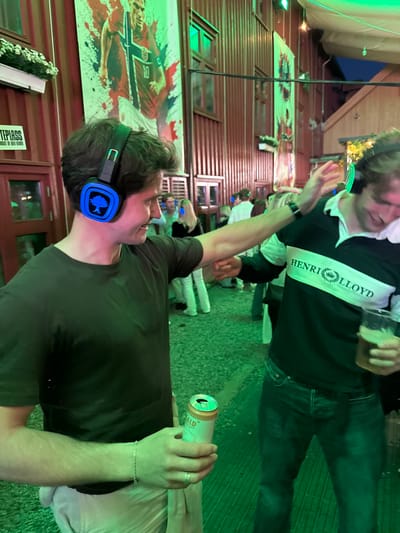 A group of people are gathered outdoors in what appears to be a lively event or party, with a strong green lighting illuminating the scene. In the foreground, a man wearing blue headphones with a tree logo is smiling and looking to his right, holding a can in his right hand. He is dressed in a dark olive t-shirt and light-colored pants, with a silver bracelet on his left wrist and a ring on his right hand. To his right, another man in a dark green and white striped rugby shirt with "HENRI LLOYD" written on it is holding a glass of beer and gesturing with his left hand. In the background, other individuals are visible, some also wearing headphones, and large banners featuring a soccer player and a crest hang on a wooden-paneled building. The ground is covered with what looks like gravel or small stones. The overall ambiance suggests a social gathering possibly at a festival or outdoor venue in Tønsberg, Norway, indicated by the prominent "TEPLASS" signage which is a known venue in Tønsberg.