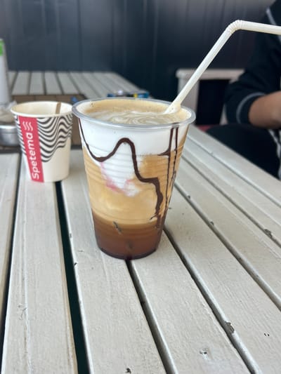 An overhead-angle image captures a clear plastic cup containing a layered iced coffee drink on a white slatted wooden table in Dolni Tsibar, Bulgaria. The drink features a dark brown base, light brown coffee, white foam with a circular pattern, and dark brown chocolate syrup drizzled inside the cup. A white, flexible plastic straw is inserted into the drink. To the left, a disposable paper cup with a "Spetema" logo in red and a black and white wavy pattern is present, containing a wooden stir stick. A small, circular metallic container is positioned between the two cups. A person wearing a dark-colored long-sleeved garment with white stripe details is partially visible in the upper right background. The background consists of a dark, solid wall or structure. The white slatted table exhibits minor wear and dirt.