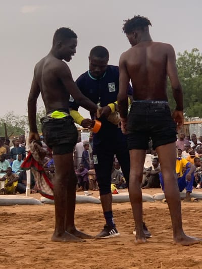 The image shows three men standing on a sandy wrestling ring. The man on the left is shirtless and wearing black shorts with a yellow waistband, holding a red and white cloth in his left hand. He is looking down at the man in the middle. The man in the middle is wearing a dark blue tracksuit with a white logo. He is wrapping something around the man on the right's right wrist. The man on the right is shirtless and wearing black shorts. His back is to the camera. The man in the middle is holding an orange object in his left hand. The three men are standing on a sandy wrestling ring. There is a crowd of people sitting in the background. The image appears to have been taken in a rural area. The coordinates 13.049, 5.237 place the image in Sokoto, Nigeria.