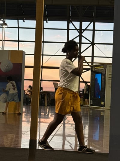 The photograph captures a person in motion, viewed from the side, walking indoors with a large window structure in the background displaying a sunset. The individual, who appears to be female with dark hair pulled back, is wearing a white t-shirt and yellow shorts, with athletic shoes. They are positioned in the mid-ground. In the left mid-ground, a display panel with large circular graphics and a young person in a white t-shirt and light-colored shorts is visible. To the right of the central figure, a large exhibit case with a blue illuminated interior shows a silhouetted figure. The environment is reflective, suggesting a polished floor. The broader context of the location is an indoor space with an extensive grid-like window system that overlooks an outdoor sky with pastel hues of orange and pink, indicative of sunset. The metallic framework of the window structure is also visible. In the far background, other individuals are dimly visible, some seated at tables. The overall scene suggests a public indoor space, possibly an airport terminal or a museum, given the exhibits and open layout. The location is specified as Zaventem, Belgium.