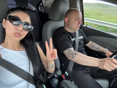 Two individuals are visible inside a vehicle. A male with a shaved head is in the driver's seat wearing a black shirt with white lettering and a seatbelt. He has tattoos on his left arm. A female is in the passenger seat wearing sunglasses and a white shirt with a seatbelt. She is holding up two fingers in a peace sign. A road and green landscape are visible outside the car window. The location is potentially near Isperihovo, Bulgaria, based on the prompt, but this cannot be confirmed from the image itself.