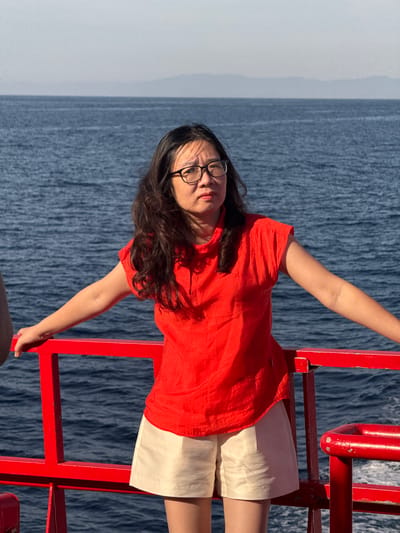 A medium shot captures a woman standing on the deck of a boat, with the ocean and a hazy horizon in the background. She is wearing glasses, a bright red top, and light-colored shorts. Her arms are outstretched, resting on a red railing. The woman's expression is serious as she looks slightly upwards and to her left. The sea is a deep blue, and the sky is a pale, overcast grey. The red railing and the woman's vibrant top provide the only strong colors in the otherwise muted scene. There is no indication of the city of Dong Son, Vietnam, in this image.