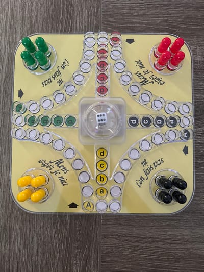 A clear plastic "Mens erger je niet" (Ludo) board game rests on a dark brown wooden plank floor. The square board has rounded corners and a yellow base. Four sets of cylindrical game pieces, each consisting of four pieces, are present in designated clear plastic holders at each corner: green in the top-left, red in the top-right, yellow in the bottom-left, and black in the bottom-right. A central clear plastic dome encloses a white six-sided die, displaying the number '6' face-up. Game tracks, composed of clear circular depressions, emanate from the central area towards each corner. Each color quadrant also features a "home" track with four marked positions (a, b, c, d) in corresponding colors, where the initial pieces are positioned. Text printed on the yellow board includes "Mens erger je niet" in Dutch and "ne t'en fais pas" in French, along with black directional arrows. The scene is located in Wanneperveen, Netherlands.