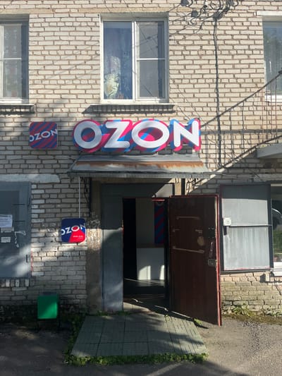 The image shows the exterior of a brick building with an open doorway. The building has a weathered appearance, with some discolored bricks and peeling paint. Two windows are visible on the upper left side of the building. A large, illuminated sign with the letters "OZON" in white with blue and pink outlines is prominently displayed on the left side of the building. Below this, two smaller square signs also bearing the "OZON" logo are visible. One of these smaller signs has the text "09:00-21:00" printed on it. The doorway to the building is open, revealing a dark interior. The ground outside the building is a mix of paved tiles and asphalt, with some green foliage growing along the edges. The lighting suggests it is daytime, with shadows cast by the building and the open door. The scene appears to be an entrance to a retail or service location in Серебрянский, Russia, indicated by the presence of the OZON branding. There are no people visible in the image.