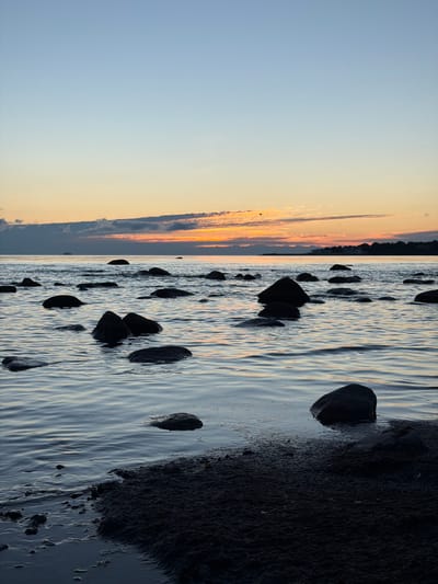 This image depicts a serene sunset over the Baltic Sea in Haabneeme alevik, Estonia. The sky transitions from a pale blue at the top to soft oranges and pinks near the horizon, where scattered clouds are illuminated by the setting sun. A distant ship is visible on the water. The foreground is dominated by a calm sea with numerous dark, rounded rocks emerging from the water and along the sandy shoreline. The water exhibits gentle ripples, reflecting the muted colors of the sky. The lighting suggests the scene is captured during the twilight hours. There are no people or visible actions taking place.