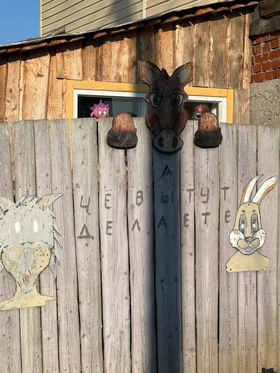 The image depicts a weathered wooden fence in Kondas, Russia, adorned with several whimsical elements. A light-colored wooden fence, with vertical planks, forms the primary structure. Attached to the fence are cutout wooden figures of animals: a grey wolf-like creature on the left, a horse's head centrally positioned above the fence, and a rabbit on the right. The horse's head is positioned as if peeking over the fence. Black lettering is painted vertically on the fence, spelling out "ЧЁ ВЫ ДЕЛАЕТЕ" which translates to "WHAT ARE YOU DOING." Above the fence, a portion of a wooden building with a window is visible. A pink, cartoonish stuffed creature with large eyes is hanging inside the window. The overall scene suggests an outdoor, rural setting.