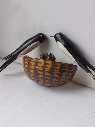 This is a close-up, eye-level, still photograph of a decorative bird's nest with three figurines of baby birds inside, and two adult bird figurines perched on the edge. The nest is made of wood and has a woven basket texture, with a dark brown, almost black, woven pattern on a lighter brown background. The adult birds are black with white undersides and white eyes. They appear to be swallows, with long, forked tails. One adult bird is on the left side of the nest, facing towards it, and the other is on the right side, also facing towards the nest. The baby birds are dark brown or black, with their mouths open, likely awaiting food. The scene is set against a plain white background, suggesting an indoor environment or a well-lit surface. There are no visible signs of the city of Melioratyvne, Ukraine. The overall impression is of a charming, handcrafted decorative item.