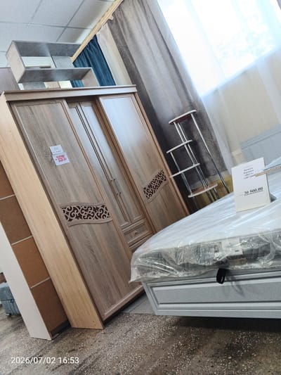 This is an indoor photograph taken at a furniture store, likely in Chany, Russia, as indicated by the timestamp. The image features a bedroom furniture display. In the foreground, a large, bed is covered in clear plastic packaging, with its base partially visible. A price tag with the text "Кровать Парящая «ВИТА» 1400*2000" and "30 500.00" is prominently displayed on the bed. To the left of the bed is a tall wooden wardrobe with multiple doors and decorative carvings. A red and white tag with "-15%" is attached to one of the wardrobe doors, suggesting a sale. Further to the left, a light-colored shelving unit is mounted on the wall. In the background, grey and light-colored curtains hang near a window, allowing bright natural light to illuminate the scene. A slender, multi-tiered metal display stand with wooden shelves is positioned between the wardrobe and the bed. The overall setting appears to be a retail environment, with the furniture arranged for display. The lighting suggests it could be daytime. There are no people visible in the image.