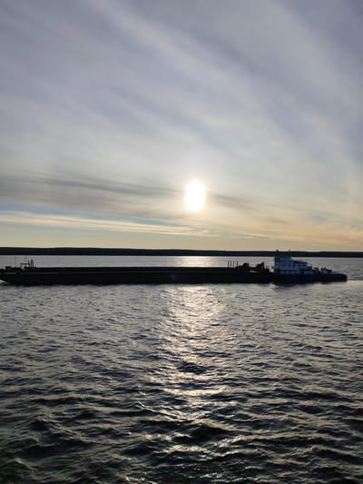 A long, flat barge is on the water under a sky where the sun is visible. At the end of the barge is a tugboat. The location is Shchetinskoe, Russia. The water is rippled, and the sun's reflection creates a shimmering effect on its surface. The horizon shows a dark line of land. The sky has light clouds with a hint of yellow from the sun.
