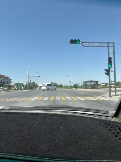 The image shows a view from inside a vehicle, looking out at a street scene. The dominant colors are blue from the sky and grey from the road and traffic signals.
The main subjects are traffic lights, a road with pedestrian crossings, and a few vehicles. There are two sets of traffic lights visible. The one on the left has a green light illuminated, and the one further down the road also appears to have a green light. Several vehicles are present, including a white van or bus and possibly other cars, though they are out of focus. The road has clear yellow and white striped pedestrian crossings. In the background, there are buildings and trees, suggesting an urban or semi-urban environment. A street lamp is also visible.
The image appears to capture a moment during daytime, with a clear blue sky and bright sunlight, indicating fair weather. The presence of traffic lights and vehicles suggests that this is a street or intersection where traffic is regulated. The perspective from inside the car indicates that the vehicle is either stopped at a red light or moving slowly through the intersection.
There is no visible text in the image. The location context of Pakhtakor, Uzbekistan, provides additional information about the setting.