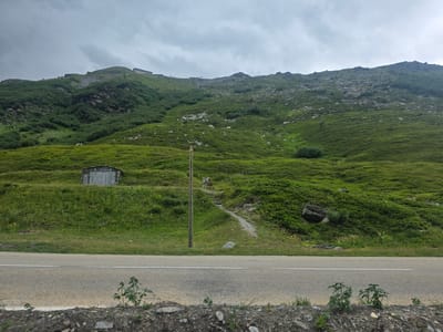 The image shows a mountainous landscape with a road in the foreground. The mountain slopes are covered with green vegetation and rocks. A small, rustic building with a memorial plaque is visible on the slope. A wooden pole with a yellow marker stands next to a path leading up the mountain. The sky is overcast. The image appears to be taken in the region of Séez, France.