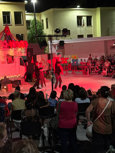 The image depicts an outdoor circus or theatrical performance at night in an urban setting, specifically in Parabita, Italy. The scene is illuminated by artificial lights, including stage lighting that casts a red glow over the performance area.

In the foreground, an audience of approximately 50-70 individuals, varying in age, is seated on dark chairs or standing. Several wheelchairs are visible among the audience members on the left and right sides. The audience faces the stage, with some individuals obscured from view due to the perspective.

The midground features a circular performance area outlined by red light. Three male performers are active on this stage. On the far left, a performer in a dark suit and fedora is interacting with another performer in the center, who wears patterned trousers and possibly a hat. The third performer, on the right, is dressed in dark clothing, holding a red piece of fabric, and appears to be balancing on a unicycle. Behind them, a white recreational vehicle (RV) acts as a backdrop on the left, decorated with colorful ribbons, red paper lanterns, and string lights. Adjacent to the RV is a tall, black rectangular panel. A banner behind the performers displays the text "NOTTE DE CIRCO" (Night of Circus). Further back, colorful, stylized flower cutouts in red, yellow, and blue, along with white balloons, are arranged behind a red barrier system. The stage is lit by multiple overhead stage lights mounted on a truss structure.

In the background, two light-colored, multi-story buildings with multiple dark windows are visible under general street lighting. A prominent green tree stands between the two buildings. An Italian flag hangs vertically from a window on the right-hand building, corroborating the stated location.