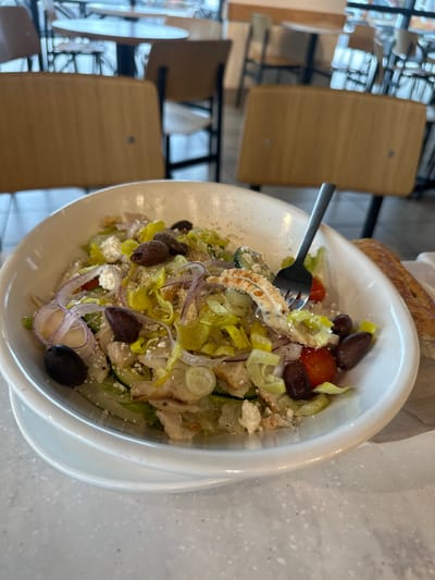 The scene, located in Medford, United States, presents a detailed view of a large, white ceramic bowl containing a chicken Greek salad, resting on a light gray speckled countertop. The salad's components include shredded green lettuce, diced grilled chicken, thin slices of red onion, numerous dark purple Kalamata olives, bright yellow-green banana pepper rings, red cherry tomato halves, and crumbled white feta cheese. A silver-colored metal fork is embedded vertically into the salad, with pieces of chicken and lettuce visible on its tines. To the right of the bowl, a segment of a golden-brown breadstick is placed on a white paper liner or napkin. The background is an out-of-focus indoor restaurant environment, revealing several light-toned wooden chairs with dark metal frames and round tables, suggesting a dining area.