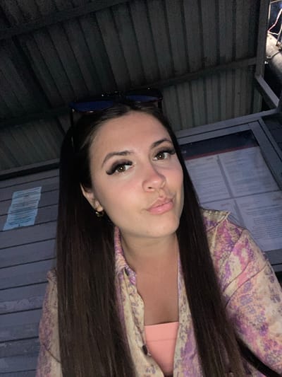 In Samur, Russia, a young woman with long, dark hair and dramatic false eyelashes is pictured in a close-up selfie. She is wearing a colorful patterned shirt over a pink garment, and a pair of sunglasses rests on her head. The background appears to be an outdoor covered area, possibly a bus stop or waiting shelter, with corrugated metal roofing and siding. A notice board or informational display is visible behind her. The lighting suggests it is nighttime or a dimly lit environment, with some light sources creating highlights on her face and the environment.