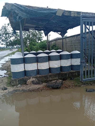 In Igrita, Nigeria, a row of approximately 10-12 cylindrical containers, painted with alternating grey and white horizontal bands, are stacked on a concrete platform. The containers appear to be newly painted or in good condition. A dilapidated metal awning covers the containers. To the right of the containers is a gray metal gate. Behind the containers, a grey cinderblock wall is visible. A flooded area with murky brown water is in the foreground, reflecting the containers and the overcast sky. A paved road with vegetation on either side is visible in the background, along with a car traveling on the road. The scene appears to be outdoors, possibly near an industrial or storage area. There are no visible people interacting with the objects. There are some rocks along the edge of the standing water.