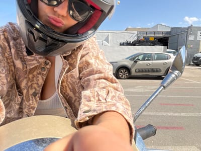 The image captures a selfie-style, low-angle perspective of a person on what appears to be a motorcycle or scooter, set against an outdoor urban or industrial backdrop under bright daylight.

The main subject is a person partially visible in the foreground, wearing a black motorcycle helmet with red interior padding and dark sunglasses. Their lips, tinted pink, are visible below the helmet visor. They are wearing a light brown or beige patterned jacket with buttons, possibly a denim-like fabric, over a ribbed, light-colored (likely white or cream) tank top or undershirt. A bare arm or forearm is also visible, positioned near the vehicle's controls.

Parts of the motorcycle/scooter are prominent: a chrome handlebar or mirror stem extends into the frame, and a black rubber grip with a textured pattern is visible, faintly displaying the brand name "Vespa".

In the mid-ground and background, the scene reveals an asphalt surface with painted red and white lines, suggesting a road or parking area. A grey modern car, resembling a Citroën C3 Aircross with black alloy wheels, is parked. Beyond the car, various industrial or commercial buildings with grey walls and some corrugated metal siding are present under a clear blue sky with scattered white clouds. A stack of black plastic crates is visible near one of the buildings. Small signs are present on the buildings, but their text is unreadable. The overall setting suggests a sunny day in an urban or industrial environment.