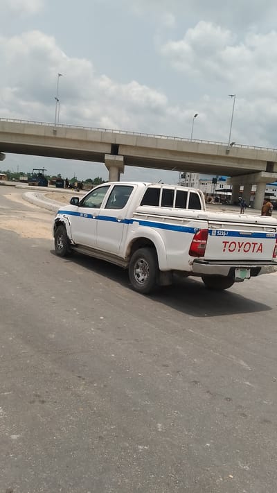 The image shows a white Toyota pickup truck with blue stripes parked on a paved road under a concrete overpass in Effurun, Nigeria. The truck has the registration number "B 5235" displayed. The front passenger side of the truck appears damaged. In the background, construction work is underway, with a tractor and a group of people visible. Two people in uniform stand nearby. The sky is overcast.