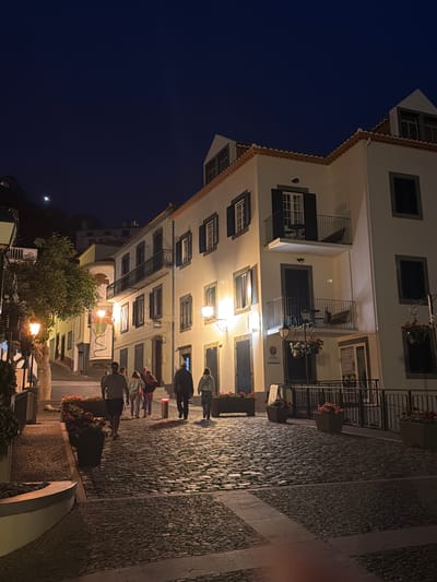 The image is a view of Ponta do Sol, Portugal, at night. The sky is dark blue. A multi-story building, likely a hotel or apartment complex, is on the left. It has balconies and numerous windows. People are walking on the stone-paved street in front of the building. Streetlights illuminate the scene. Trees and vegetation are at the bottom of the image.